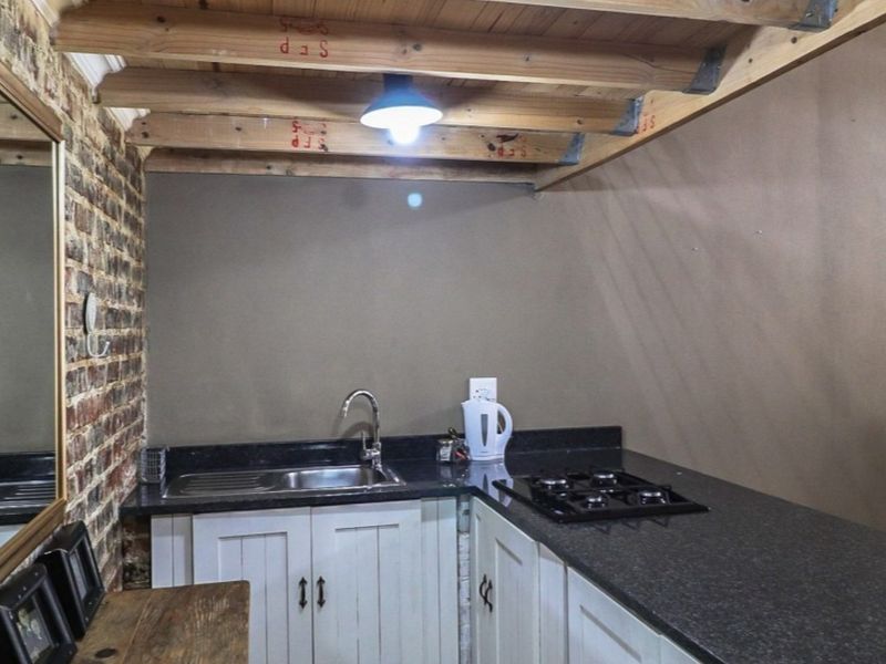 Compact kitchen featuring exposed timber ceiling beams, rustic brick wall, granite countertops, gas hob, and white cabinetry.