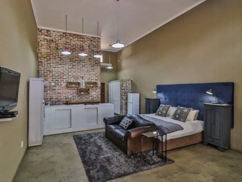 An open-plan studio apartment featuring a prominent exposed brick wall, polished concrete floors, and integrated kitchen cabinetry with modern pendant lighting.