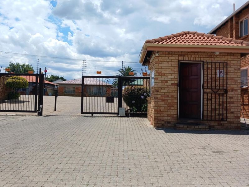 A secure townhouse complex entrance featuring a brick guard house, paved driveway, and metal security gates with electric fencing.