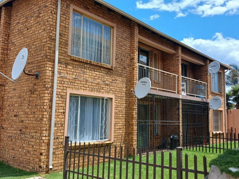 Rear view of a two-story face brick townhouse featuring a balcony, security bars on windows and doors, and perimeter metal fencing.