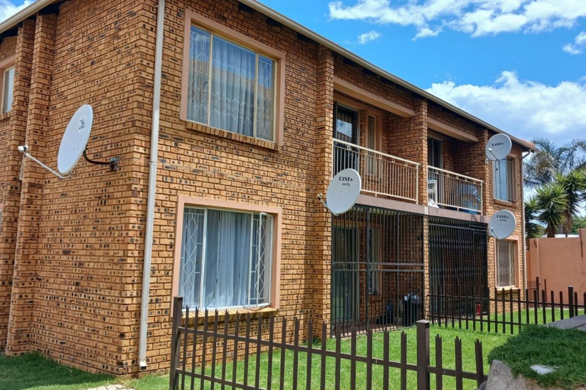 Rear view of a two-story face brick townhouse featuring a balcony, security bars on windows and doors, and perimeter metal fencing.