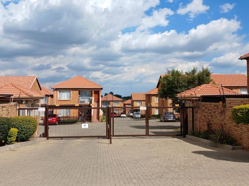 A view of the secure entrance to a townhouse complex, featuring brick buildings with tiled roofs, paved driveway, and perimeter security fencing.