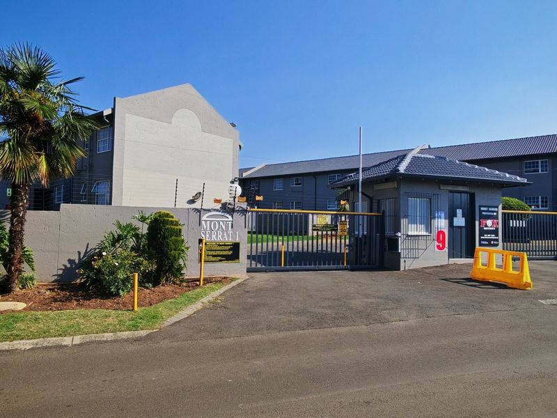 Front entrance view of Mont Serrat 1 townhouse complex featuring a secure gated access point, guard house, and perimeter wall with electric fencing.