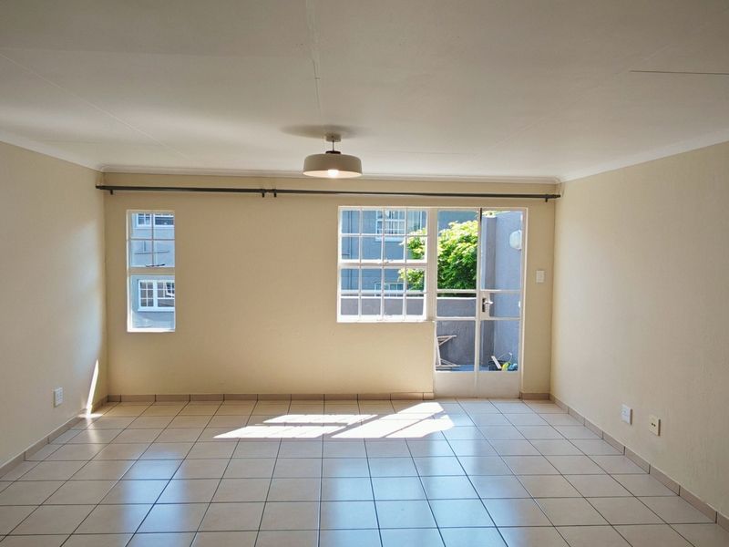 A bright, empty living room featuring neutral tiled flooring, a ceiling light fixture, and access to an outdoor area through a glass panel door.