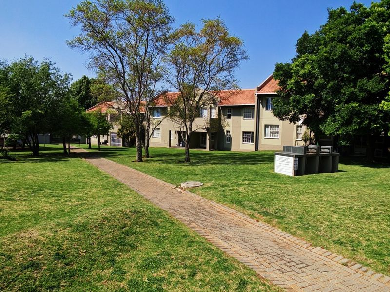 A well-maintained communal garden area featuring a paved walkway, mature trees, and a built-in braai facility set within the townhouse complex grounds.