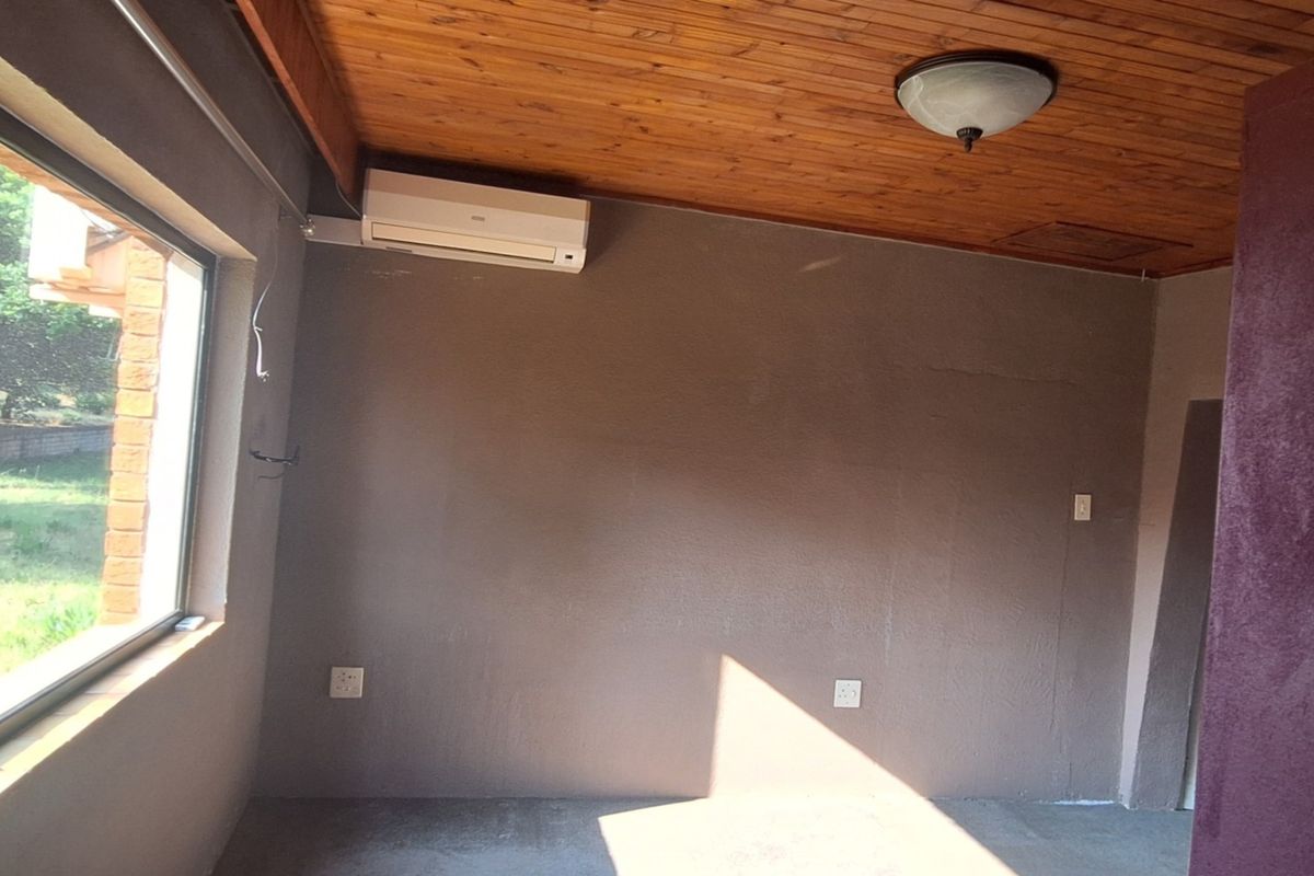 An empty living area featuring a timber ceiling, wall-mounted air conditioning unit, and large windows providing natural light and garden views.