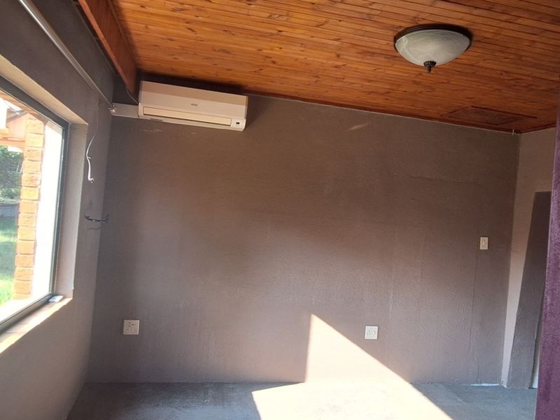 An empty living area featuring a timber ceiling, wall-mounted air conditioning unit, and large windows providing natural light and garden views.
