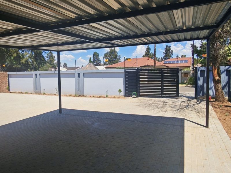 A spacious paved driveway featuring a modern metal carport structure, enclosed by a secure perimeter wall with electric fencing and a metal gate.