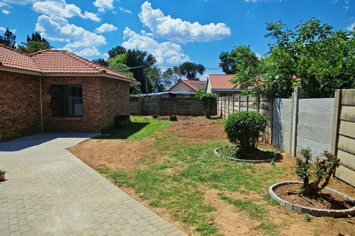 A rear exterior view of a brick house featuring a paved walkway, a precast concrete boundary wall, and a garden area with established shrubs.