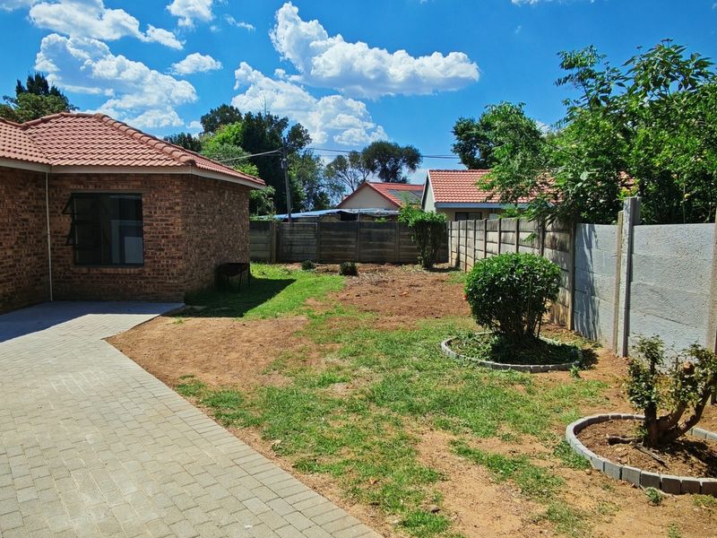 A rear exterior view of a brick house featuring a paved walkway, a precast concrete boundary wall, and a garden area with established shrubs.