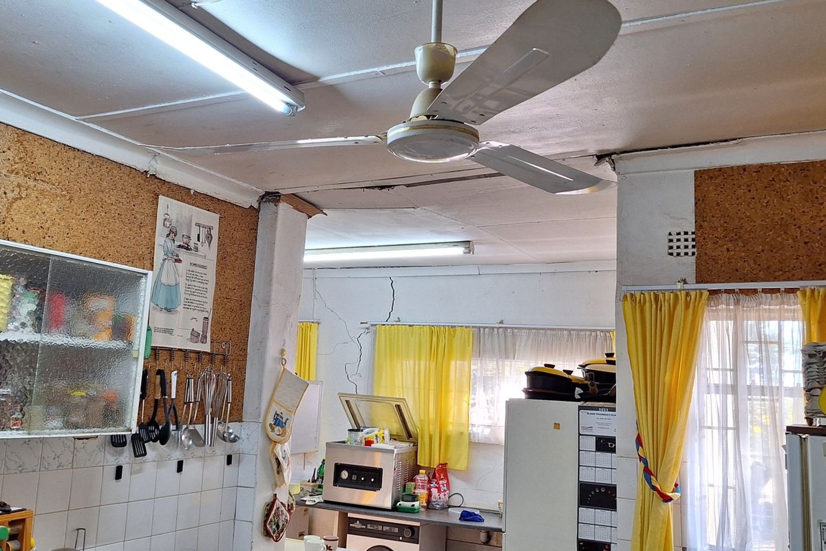 A dated kitchen featuring cork wall finishes, fluorescent lighting, and a ceiling fan, currently showing significant structural cracking on the walls and ceiling.