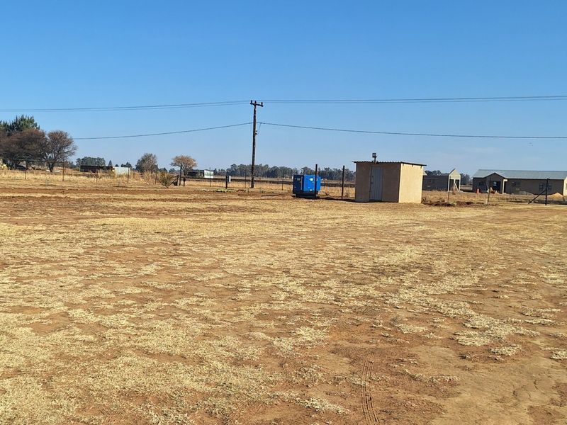 Expansive open land on a small holding featuring a utility shed, perimeter fencing, and basic infrastructure under a clear sky.
