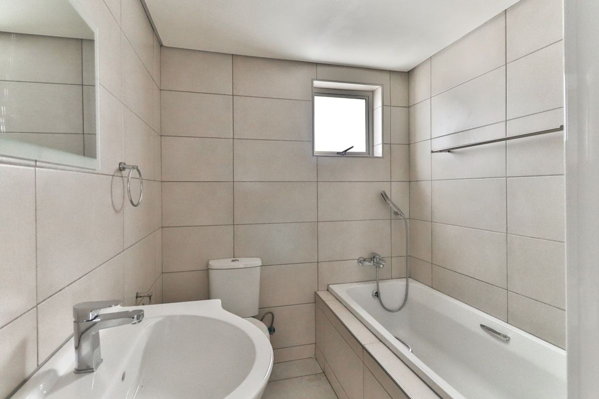 A clean, neutral-toned bathroom featuring a built-in bathtub with shower mixer, a pedestal basin, and wall-mounted towel rails.
