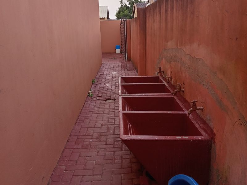 Outdoor laundry area featuring a row of built-in wash troughs with wall-mounted taps, situated along a paved side passage.
