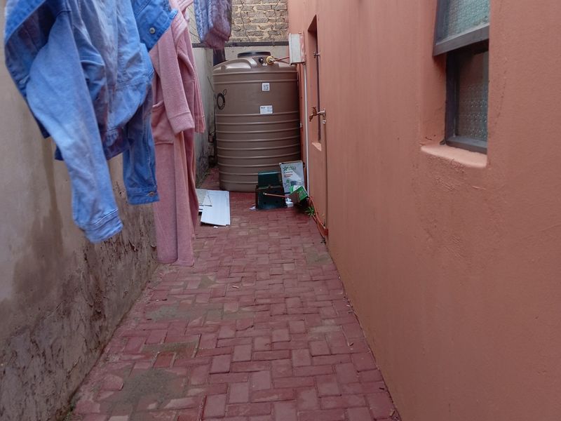 A narrow side passage featuring red brick paving, a water storage tank, and an external tap against a painted exterior wall.