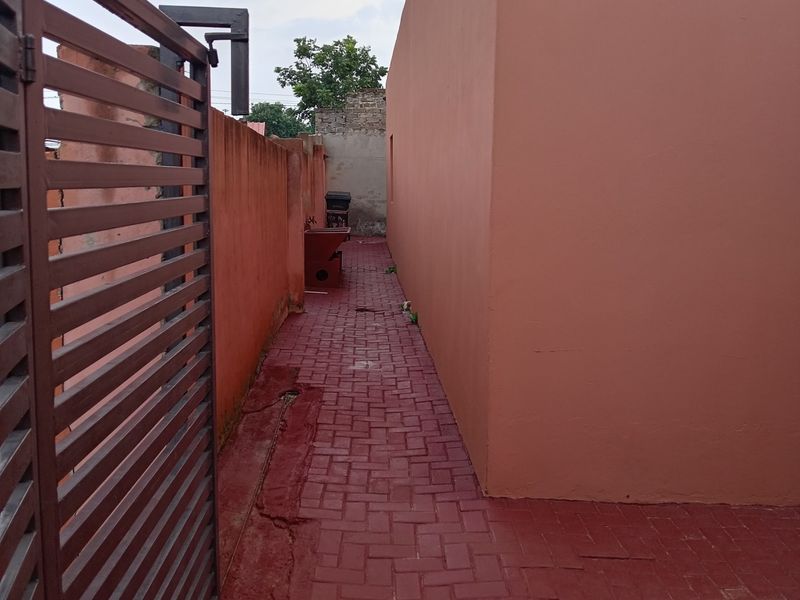 A narrow side passage featuring red painted brick paving and a metal gate, enclosed by boundary walls.