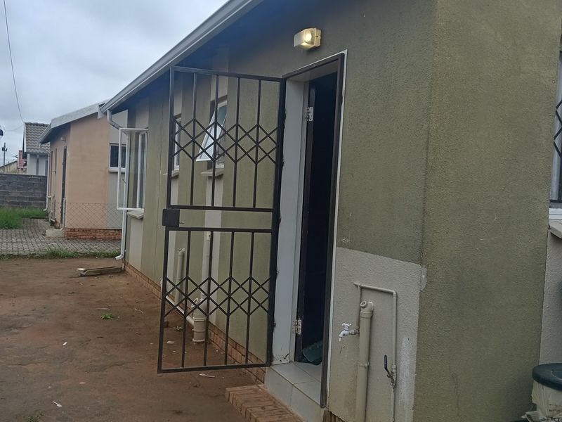 Rear exterior view of the property featuring a security gate, external lighting, and basic utility plumbing connections against a painted masonry wall.