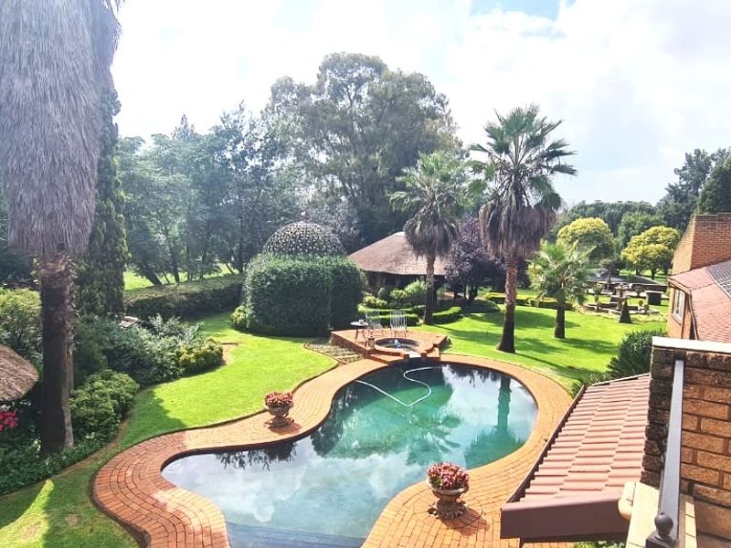 An elevated view of a landscaped garden featuring a swimming pool with brick coping, a thatched roof structure, and solar panels on the main roof.