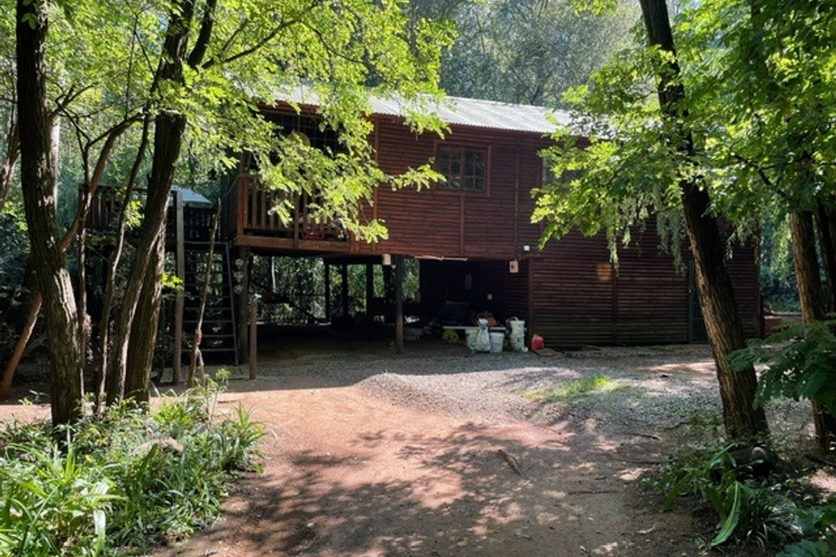 A rustic timber cabin elevated on stilts, featuring a raised deck and staircase, nestled within a wooded farm setting.