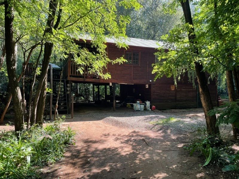 A rustic timber cabin elevated on stilts, featuring a raised deck and staircase, nestled within a wooded farm setting.