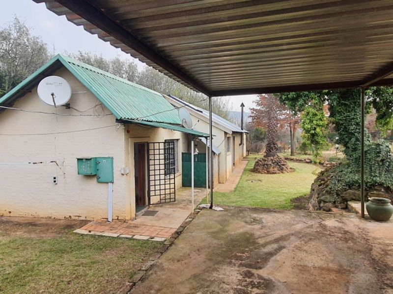 Rear view of the farmhouse featuring a covered patio area, corrugated metal roofing, and established garden surroundings with stone landscaping elements.