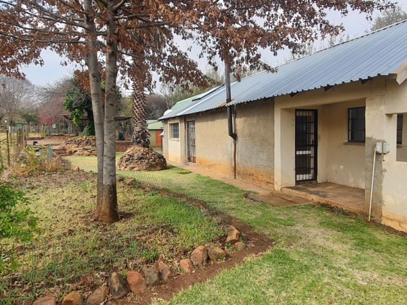 Rear view of a farm-style dwelling featuring a corrugated metal roof, plastered exterior walls, and a covered porch area overlooking the garden.