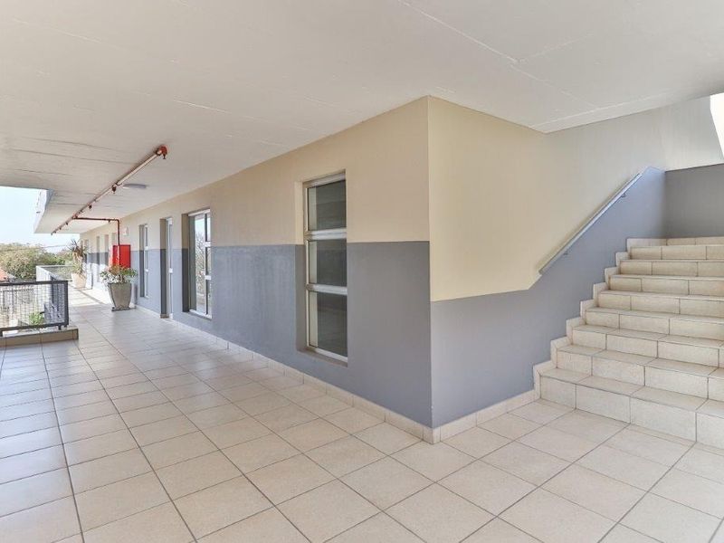 A clean, well-maintained apartment building hallway featuring tiled flooring, a staircase with a metal handrail, and exterior-facing windows.