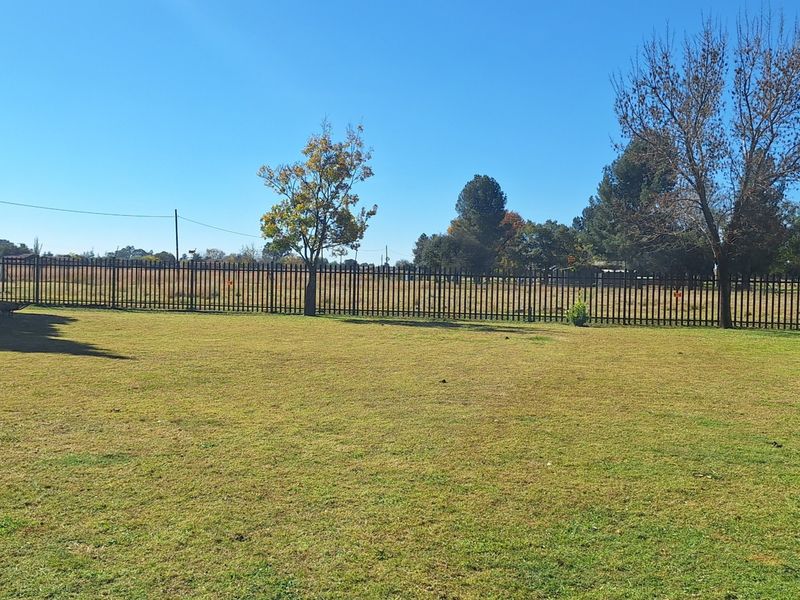 A spacious, well-maintained garden area featuring a large lawn, mature trees, and secure steel palisade perimeter fencing.