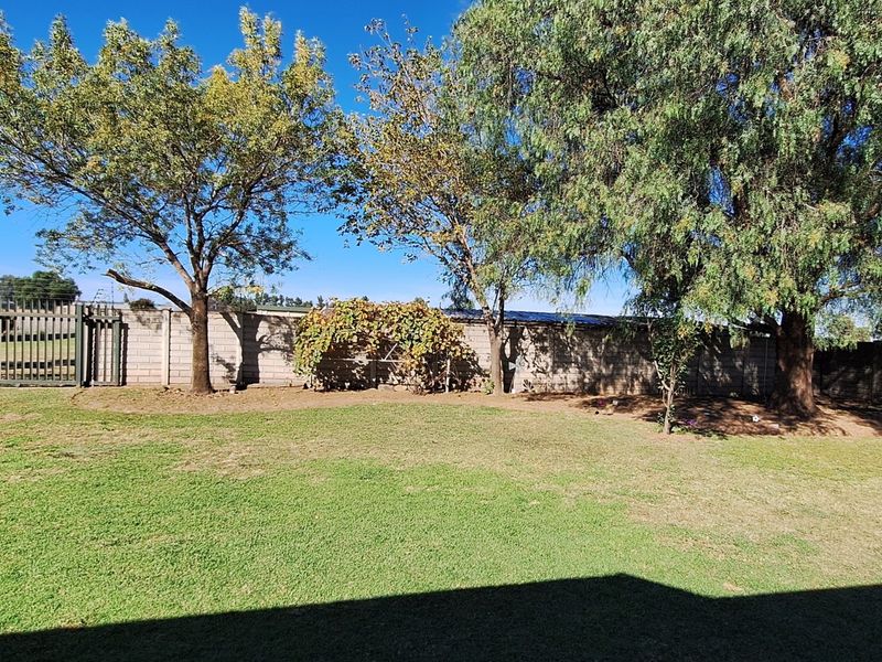 A spacious, well-maintained garden area featuring mature trees, a secure perimeter wall, and a metal entrance gate.