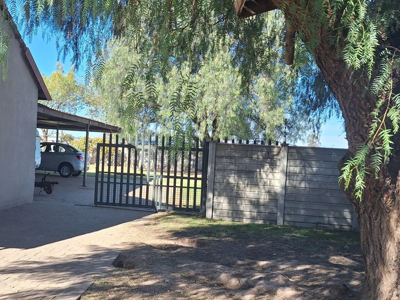 Rear exterior view showing a paved driveway, carport, metal security gate, and precast concrete boundary wall on the small holding property.