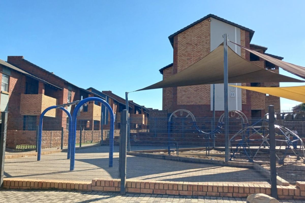 Communal outdoor playground area featuring fixed play equipment, shade sails, and secure perimeter fencing within the apartment complex grounds.