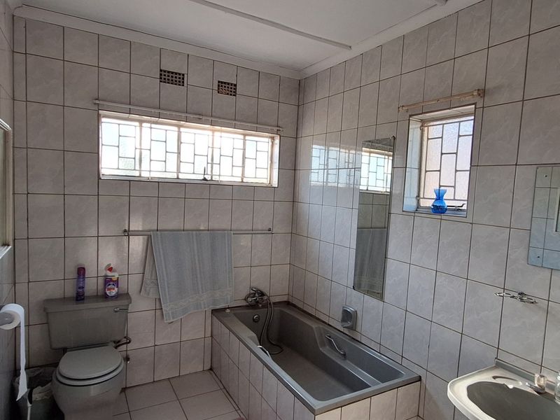 A functional bathroom featuring fully tiled walls and floors, a built-in bathtub, a pedestal basin, and a toilet with security bars on the windows.