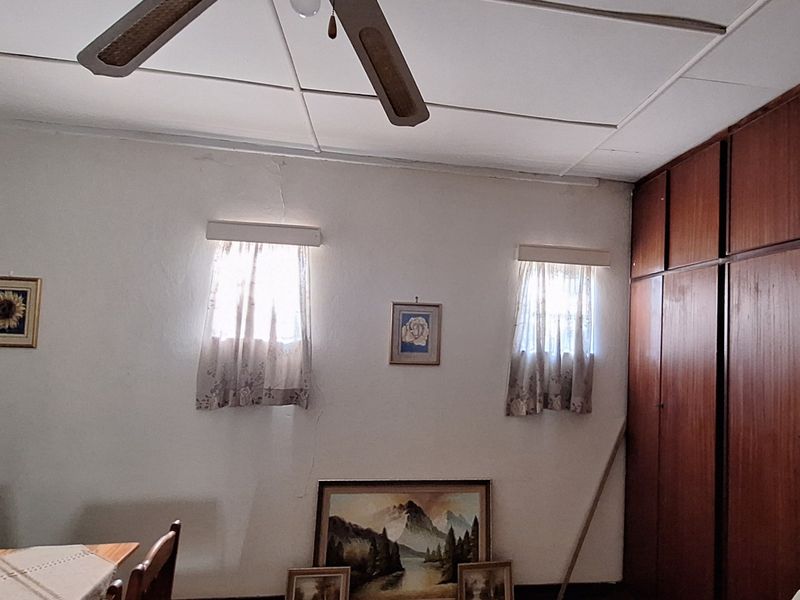 A bedroom featuring built-in wooden cupboards, a ceiling fan, and tiled ceiling, showing signs of wear including cracked wall plaster.