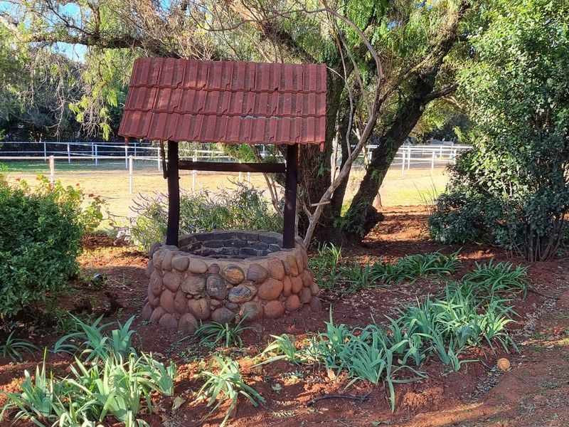 A decorative stone-clad water well feature situated within a landscaped garden area, featuring a tiled roof and surrounding mature trees and greenery.