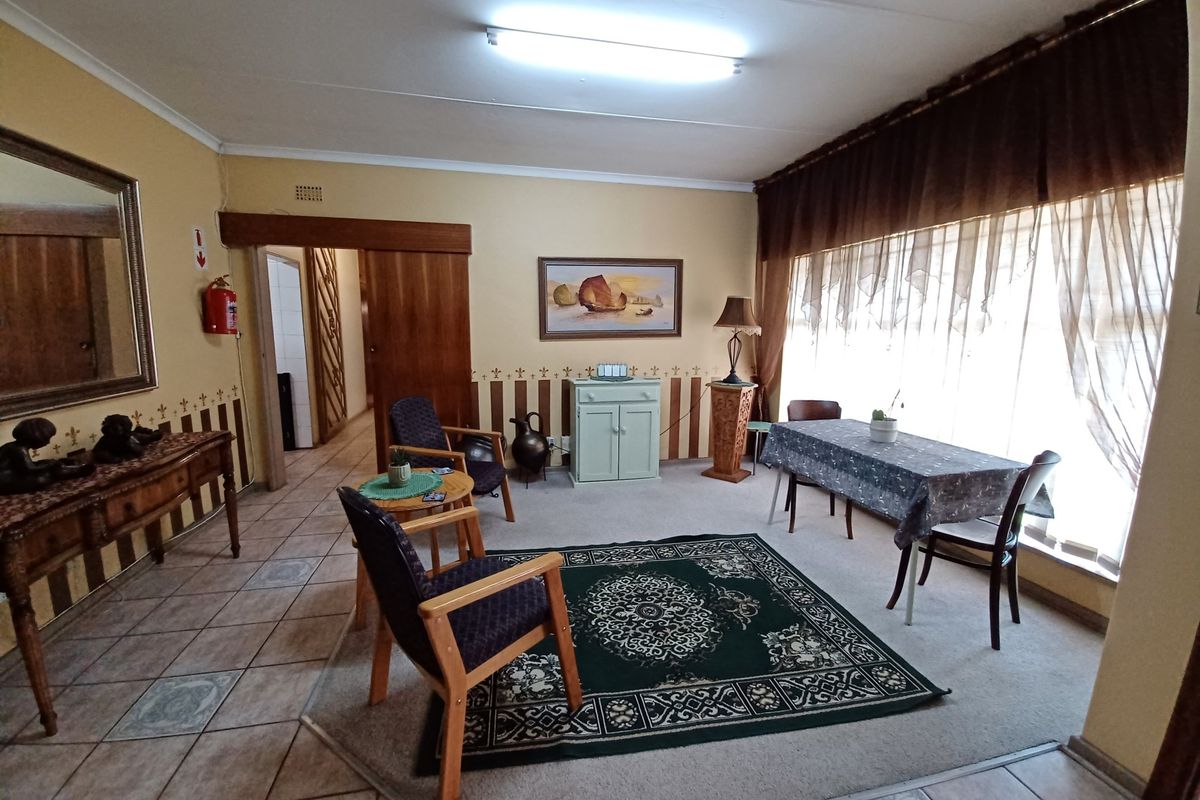 A spacious living area featuring a combination of tiled and carpeted flooring, large windows with curtains, and direct access to an adjacent hallway.