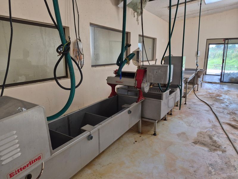 A functional utility or processing space equipped with industrial stainless steel machinery, overhead water lines, and direct access to the exterior.