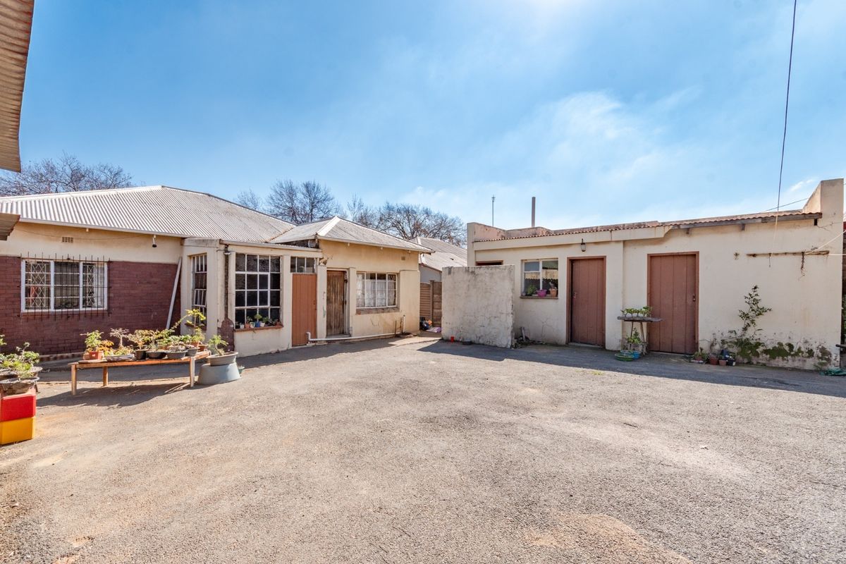 Rear exterior view of the property featuring an asphalt courtyard, multiple access doors, and original steel-framed windows with security bars.