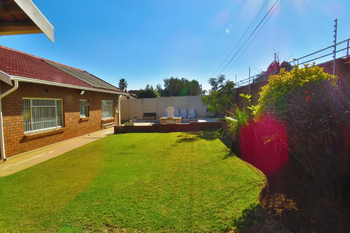 Rear garden view featuring a brick house, a well-maintained lawn, and a swimming pool area enclosed by a perimeter wall with electric fencing.