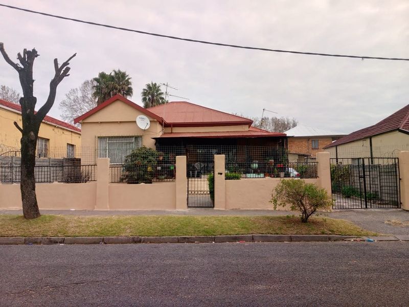 Front elevation of a single-story house featuring a corrugated metal roof, perimeter wall with security fencing, and a gated entrance.