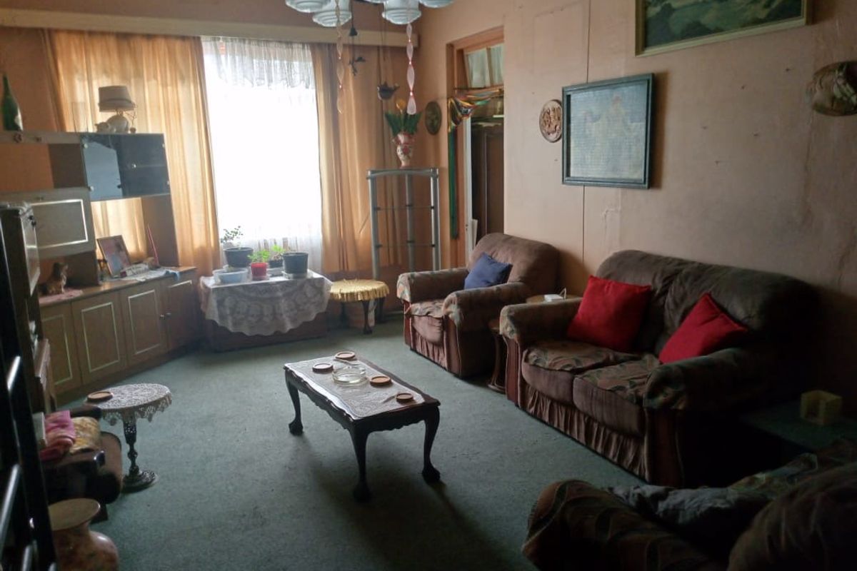 A dated living room featuring carpet flooring, a large window for natural light, and a central ceiling light fixture.