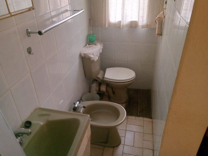A compact bathroom featuring dated sanitaryware, including a toilet, bidet, and wall-mounted basin, finished with full-height ceramic wall tiling.