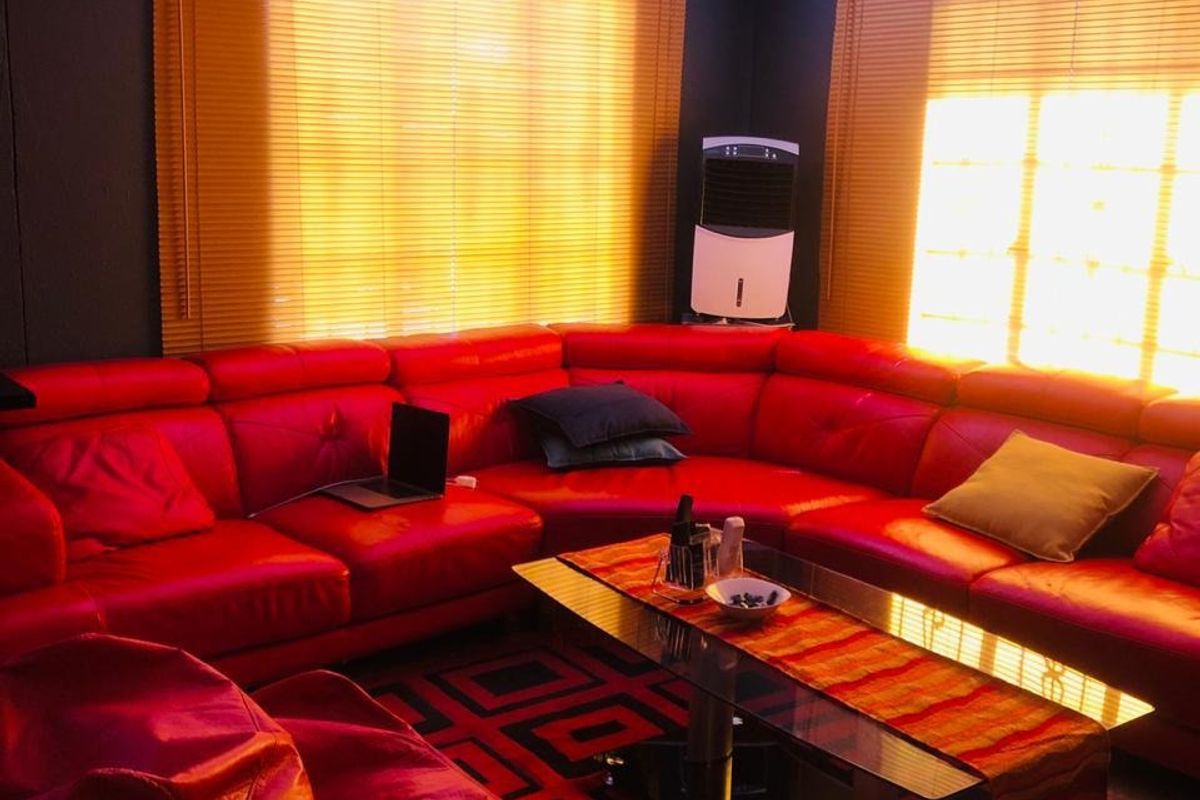 A living room featuring tiled flooring, decorative ceiling cornices, and a ceiling-mounted projector, illuminated by natural light through venetian blinds.