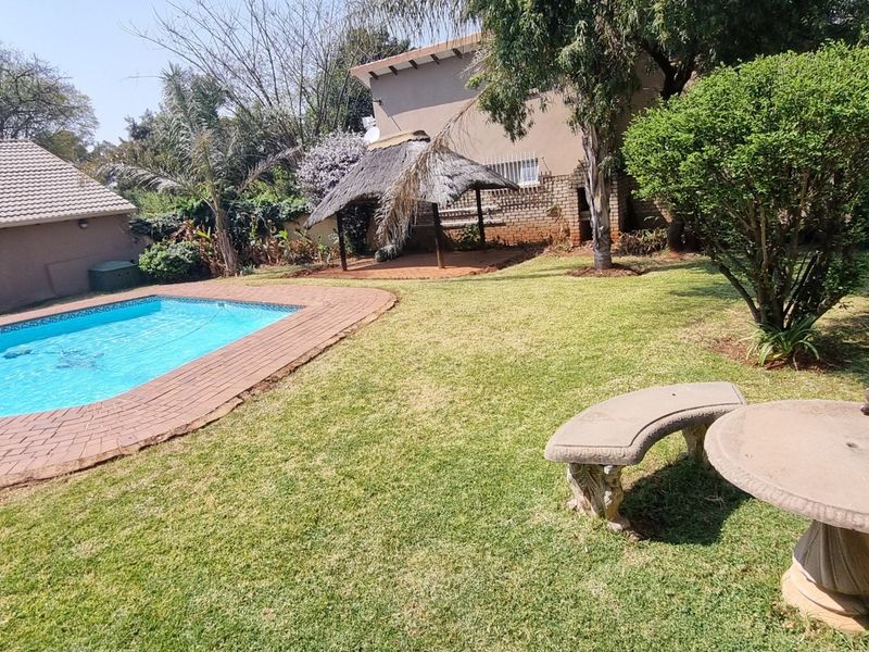 A spacious backyard featuring a swimming pool with brick coping, a traditional thatched lapa, and established garden landscaping.