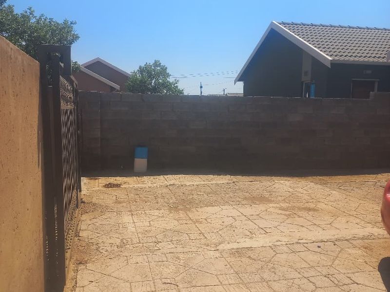 A rear exterior area featuring a paved driveway enclosed by a boundary wall and metal gate, showing signs of wear and weathering.