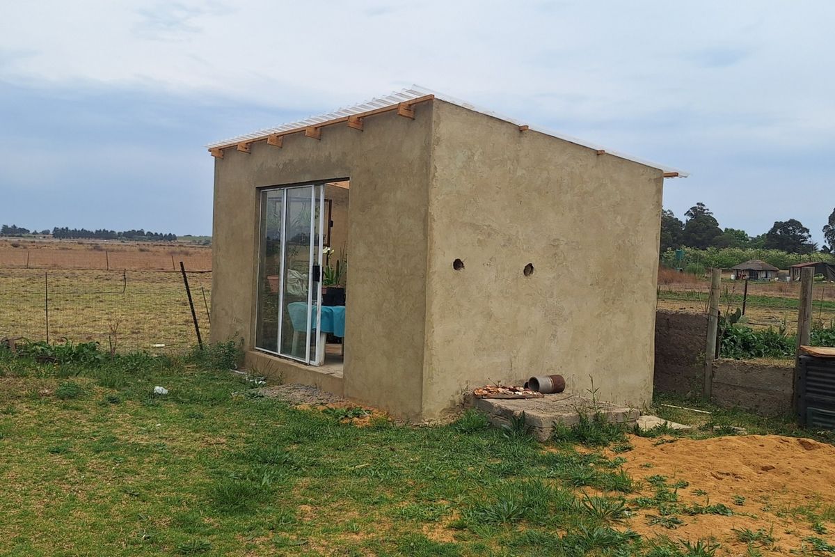 A small, detached utility structure with cement plaster walls and a sliding glass door, situated on a rural small holding.