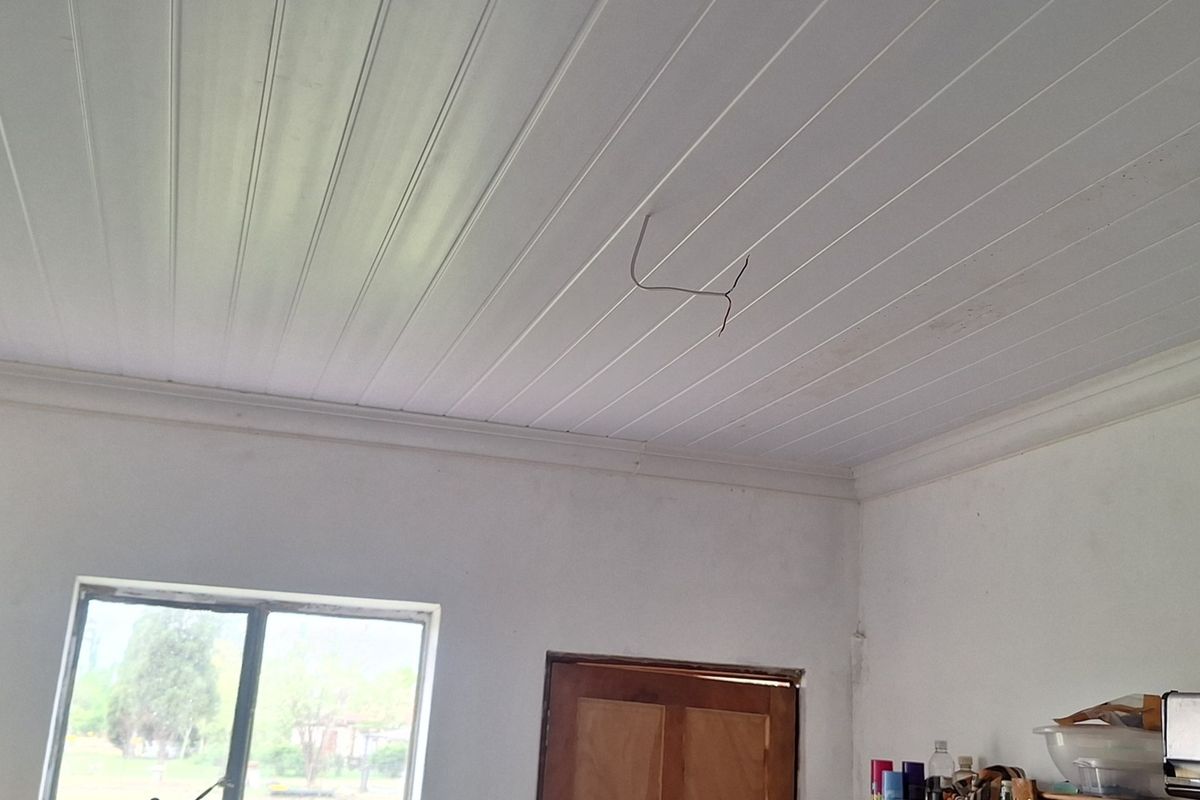 A sparsely furnished living area featuring a white tongue and groove ceiling with exposed wiring and a wooden door.