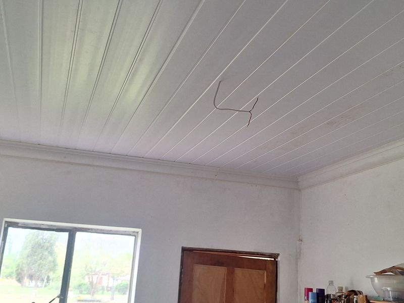 A sparsely furnished living area featuring a white tongue and groove ceiling with exposed wiring and a wooden door.