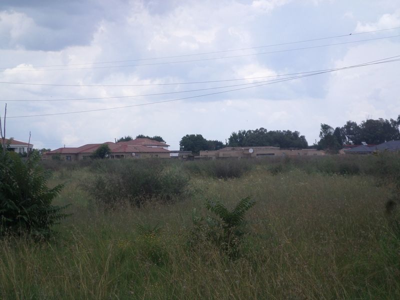 A view of an undeveloped residential plot featuring overgrown grass and surrounding neighborhood homes in the distance.