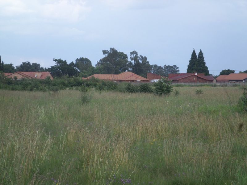 A view of a vacant residential plot featuring overgrown grass with established residential properties visible in the background.