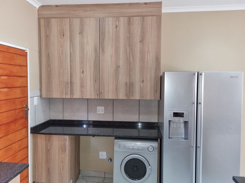 A functional scullery area featuring wood-look cabinetry, granite countertops, tiled splashback, and space for a washing machine.